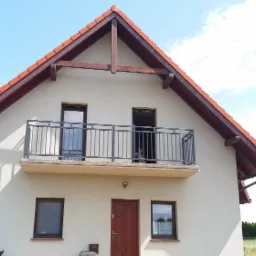 Dom jednorodzinny z balkonem zabezpieczonym szarą metalową balustradą, widok z przodu na tle błękitnego nieba.