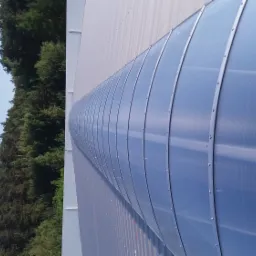 Przemysłowa konstrukcja z poliwęglanu z metalowymi wzmocnieniami w Pamiątkowie, widok z zewnątrz, na tle lasu i błękitnego nieba.