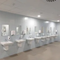 Rząd umywalek z lustrami i dozownikami w nowoczesnej, wykafelkowanej łazience publicznej oświetlonej ledami. Minimalistyczny design, jasne płytki, symetryczny układ.