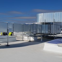 KLIMA-VENTA Sp. z o.o. Sp.k. - Instalacja wentylacyjna na dachu budynku z pracownikami w żółtych kamizelkach. Widoczne elementy systemu wentylacyjnego i świetliki dachowe.