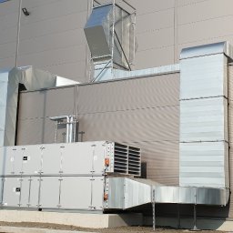 KLIMA-VENTA Sp. z o.o. Sp.k. - Zewnętrzna instalacja wentylacyjna z metalowymi kanałami na tle szarej hali. Widoczne elementy systemu i przyłącza do budynku. Profesjonalny montaż.