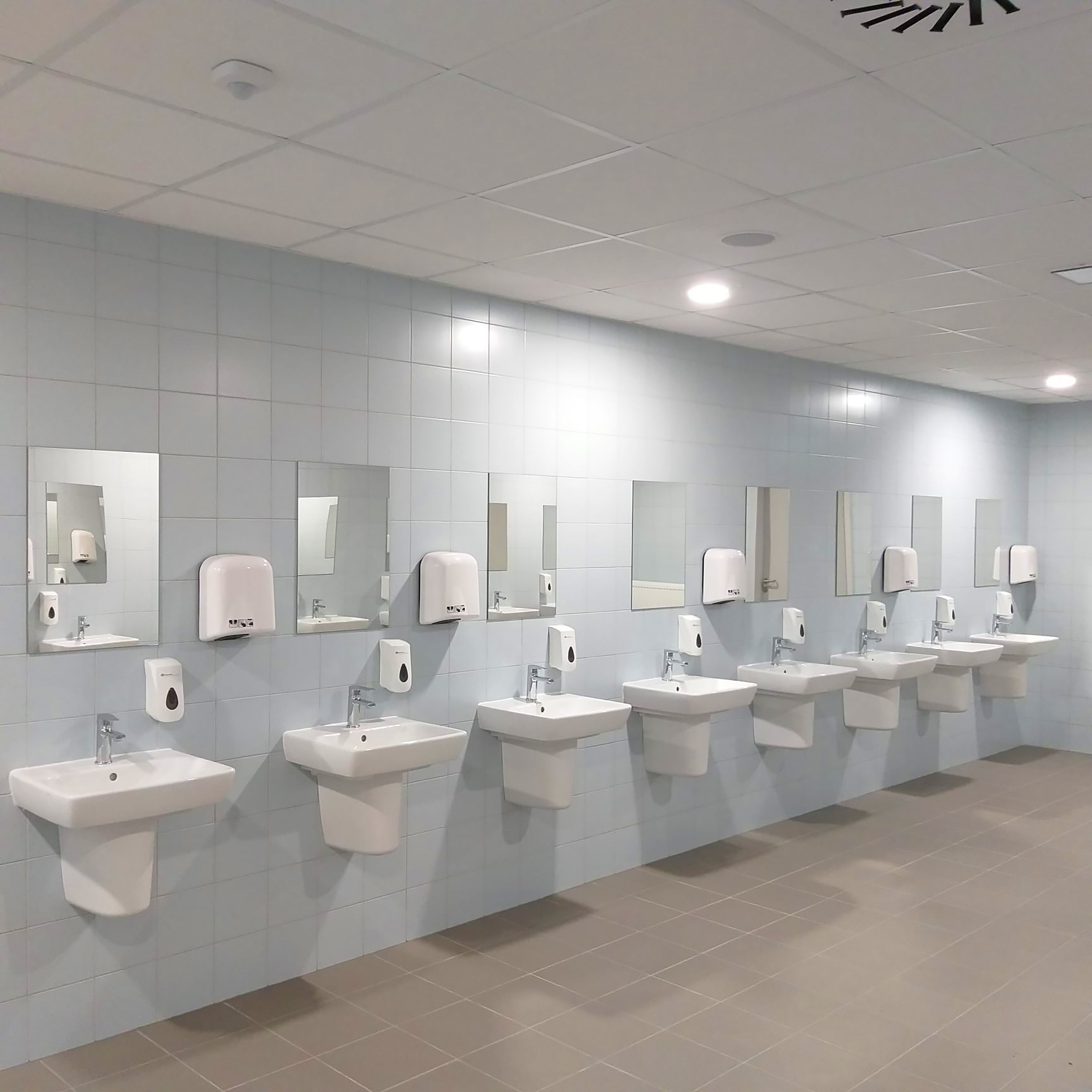 Rząd umywalek z lustrami i dozownikami w nowoczesnej, wykafelkowanej łazience publicznej oświetlonej ledami. Minimalistyczny design, jasne płytki, symetryczny układ.