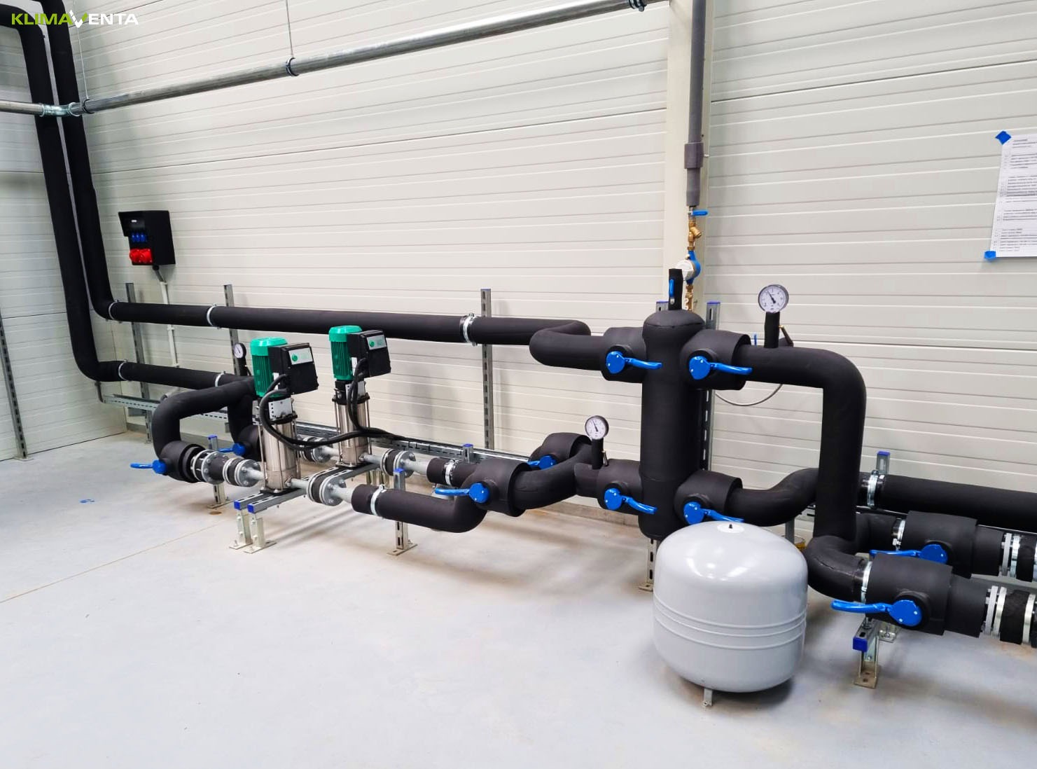 Kompleksowa instalacja grzewcza z czarnymi rurami izolowanymi, srebrnymi pompami i niebieskimi zaworami w hali z panelami ściennymi i szarą podłogą.
