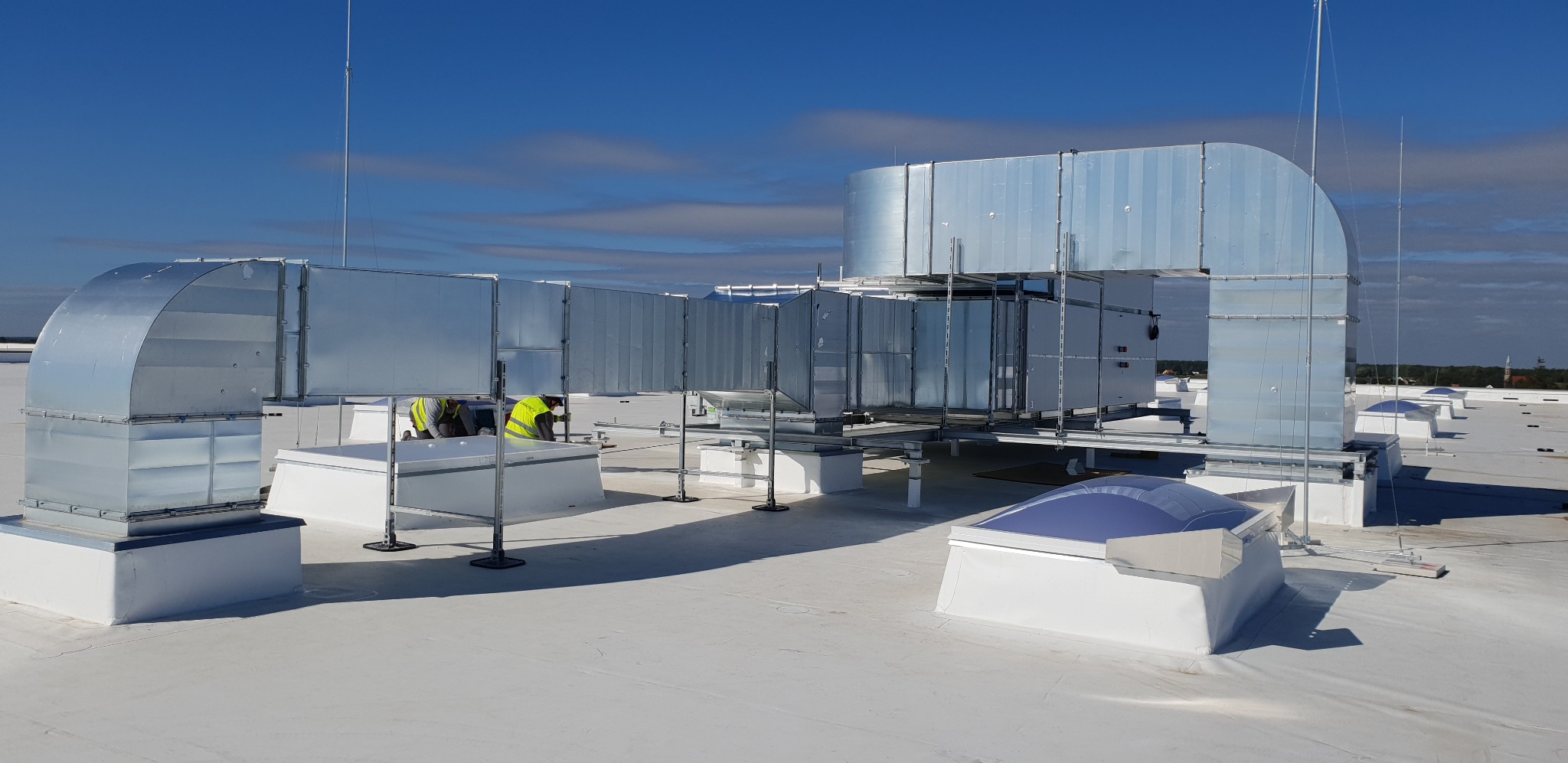 Instalacja wentylacyjna na dachu budynku z pracownikami w żółtych kamizelkach. Widoczne elementy systemu wentylacyjnego i świetliki dachowe.