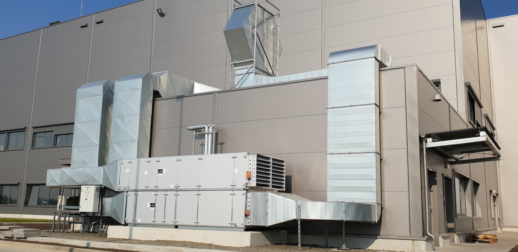 Zewnętrzna instalacja wentylacyjna z metalowymi kanałami na tle szarej hali. Widoczne elementy systemu i przyłącza do budynku. Profesjonalny montaż.