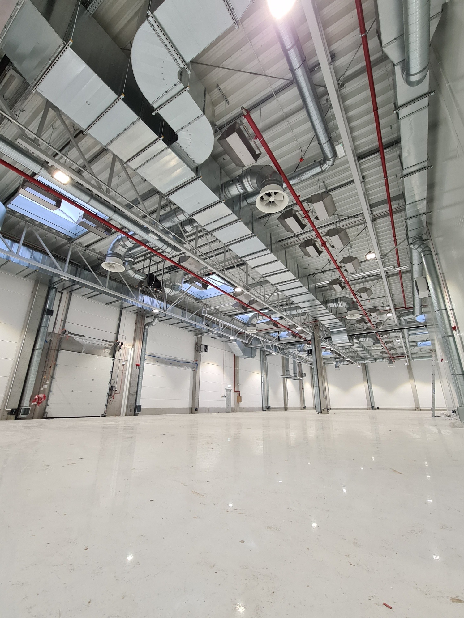 Nowoczesna hala magazynowa z systemem wentylacji i klimatyzacji pod sufitem. Widoczne rury, kanały wentylacyjne i rozproszone światło wpadające przez okna dachowe.