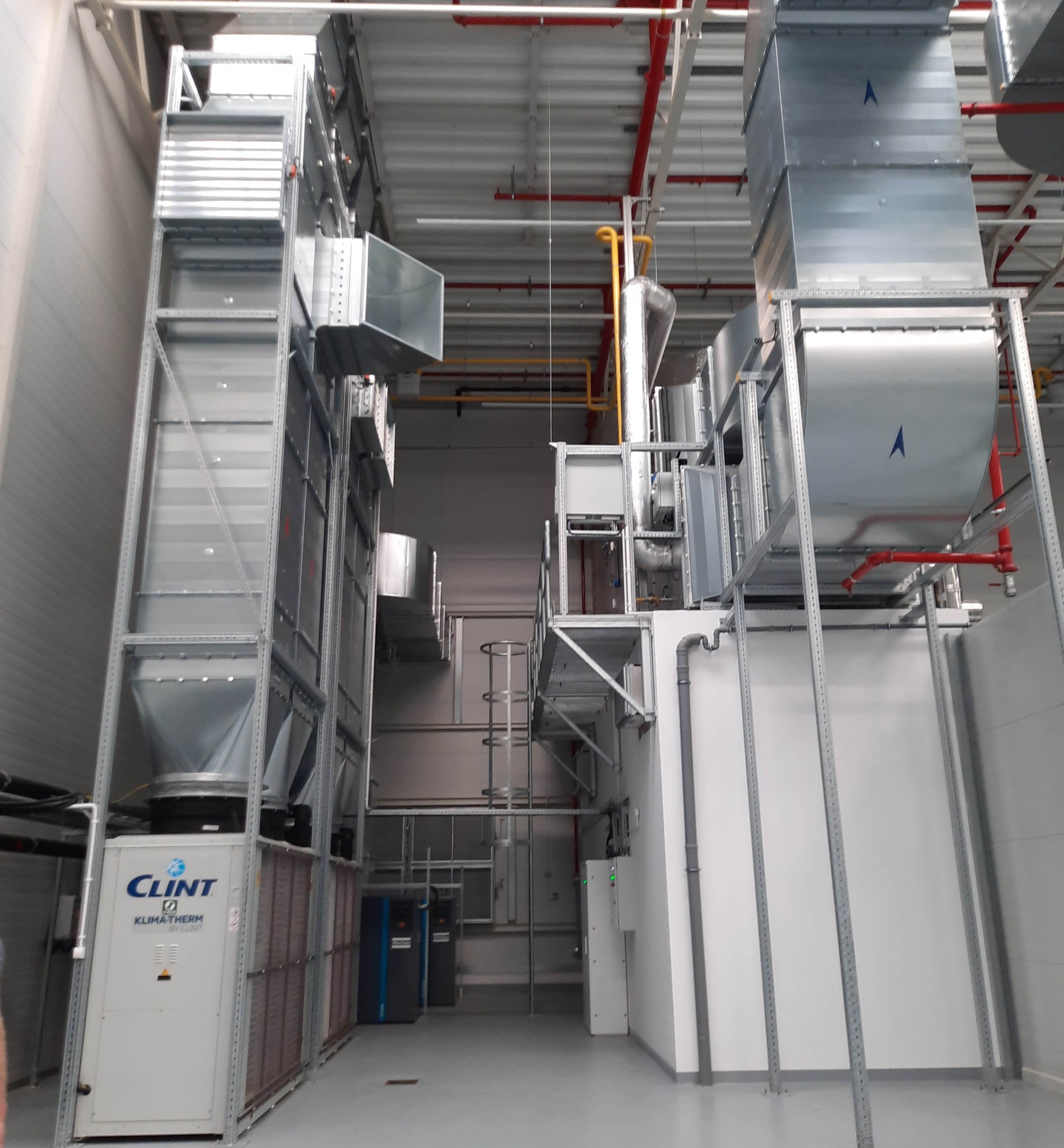 Kompleksowa instalacja przemysłowa z systemem wentylacji, rurami i urządzeniami marki Clint Klimatherm w jasnym pomieszczeniu. Widoczne elementy wsporcze i rurociągi.