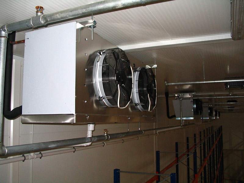 Wnętrze chłodni z zamontowanymi wentylatorami chłodzącymi i rurami instalacji, widoczne regały magazynowe.