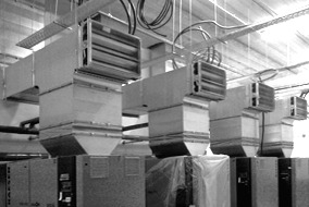 Rząd przemysłowych nagrzewnic kanałowych zawieszonych pod sufitem hali, z widocznymi przewodami i elementami montażowymi, w czarno-białej tonacji.