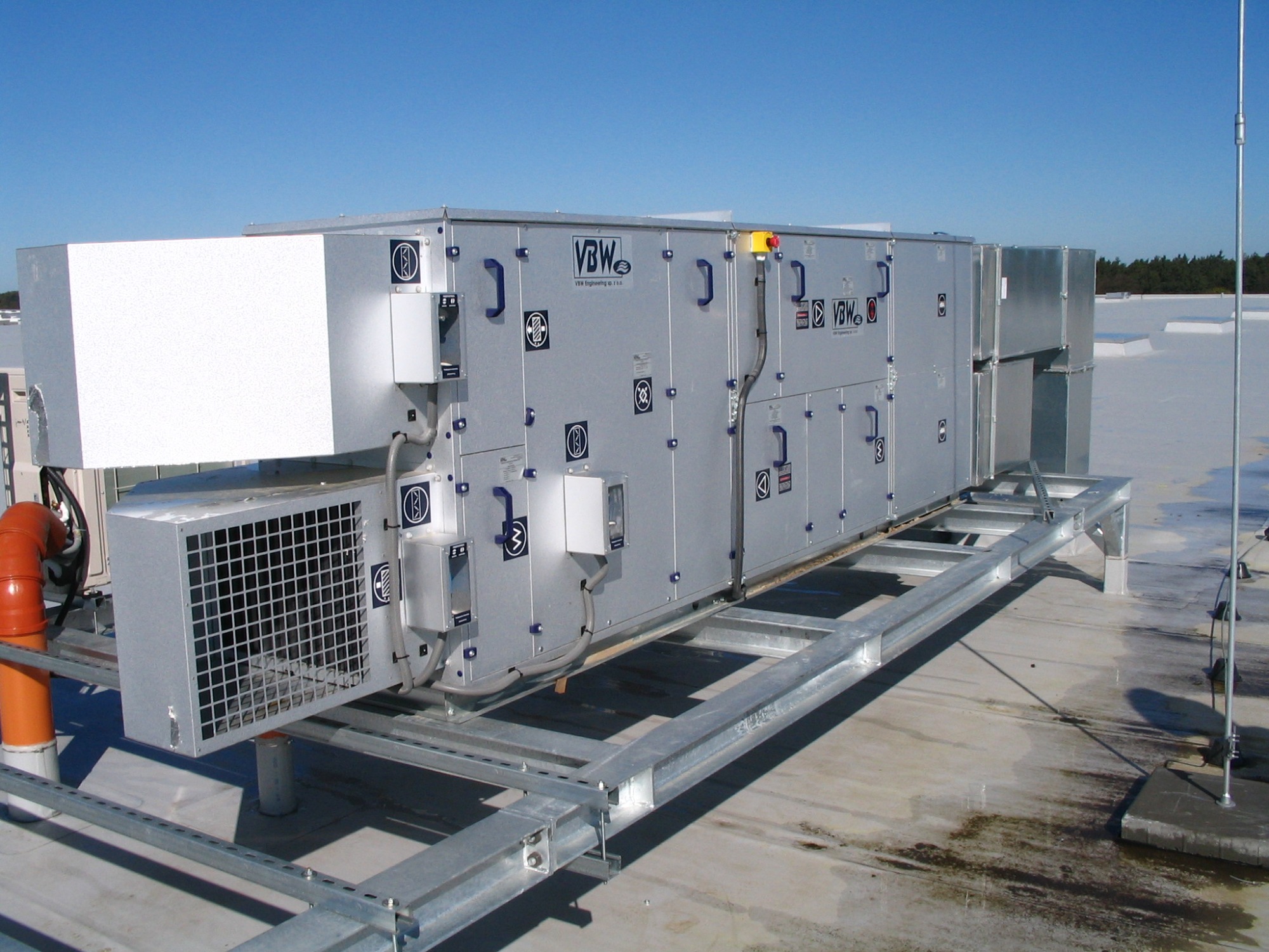 Centrala wentylacyjna VBW na dachu, widoczne logo firmy, metalowa konstrukcja wsporcza, instalacja odgromowa i fragment instalacji kanalizacyjnej z rur PCV.