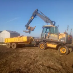 Żółta koparka kołowa Volvo EW140C z otwartą łyżką wysypuje ziemię do żółtej przyczepy na polu w słoneczny dzień. W tle widoczny budynek gospodarczy.