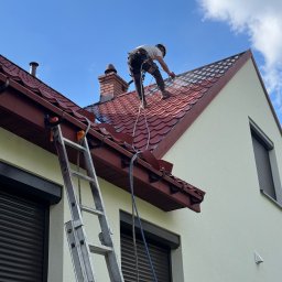 Promal-Dach - Pracownik w uprzęży, z drabiny, czyści czerwoną dachówkę domu jednorodzinnego za pomocą myjki ciśnieniowej. Widoczne zacieki i czyszczona powierzchnia.