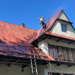 Promal-Dach - Pracownik w uprzęży, z drabiny, spryskuje wodą czerwoną dachówkę na dwuspadowym dachu domu z poddaszem, na tle błękitnego nieba. Widoczne zacieki.
