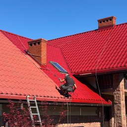 Promal-Dach - Częściowo pomalowany na czerwono dach z blachodachówki. Pracownik w uprzęży maluje dach natryskowo. Widoczna drabina oparta o rynnę i fragment elewacji domu.