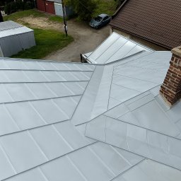 Promal-Dach - Błyszczący, srebrny dach z blachy trapezowej, widok z góry, z kominem z cegły i fragmentem otoczenia. Nowoczesne pokrycie dachowe.