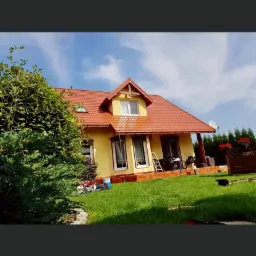 Żółty dom jednorodzinny z czerwoną dachówką, częściowo umytą elewacją i dachem, z widocznym sprzętem do mycia na trawniku.