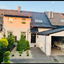 Dom jednorodzinny z ciemną dachówką i kostką brukową na podjeździe, widok z zewnątrz w słoneczny dzień.