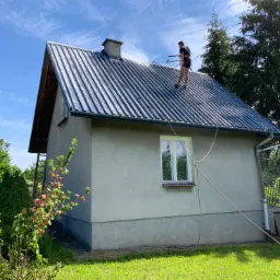 Pracownik w uprzęży czyści metalowy dach domu jednorodzinnego myjką ciśnieniową, widoczne smugi czystej powierzchni, zielony trawnik i drzewa w tle, błękitne niebo.