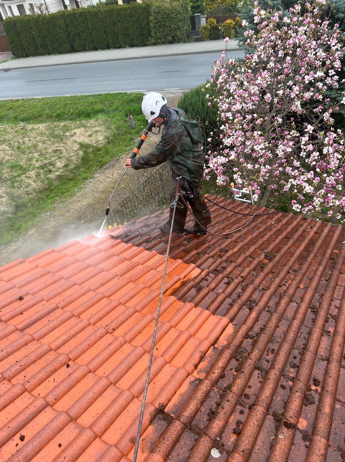 Pracownik w kasku i kombinezonie czyści dach z czerwonej dachówki za pomocą myjki ciśnieniowej. Widoczna różnica między umytą a zabrudzoną częścią dachu. W tle kwitnące drzewo.