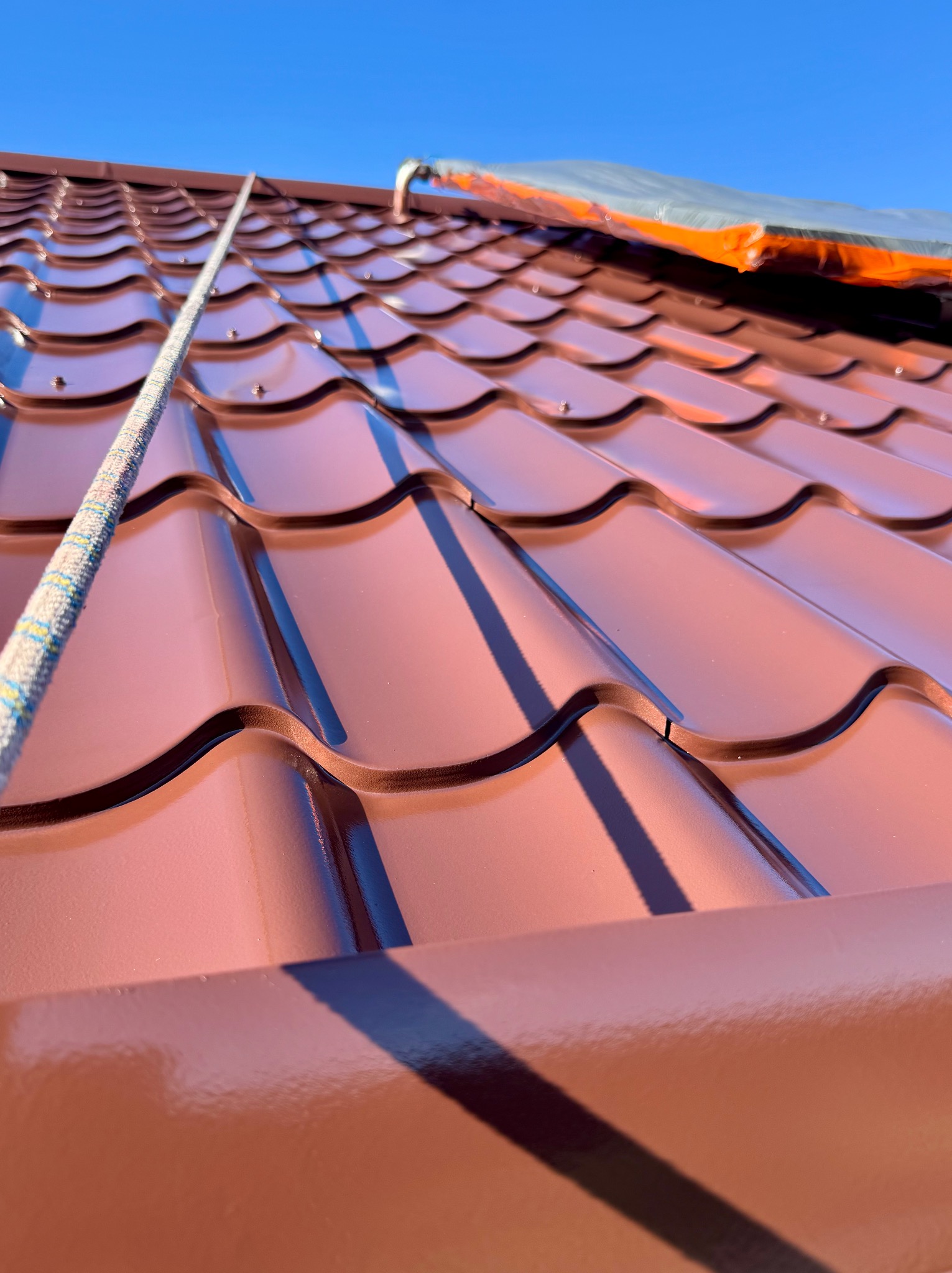 Błyszcząca, brązowa dachówka modułowa pod błękitnym niebem. Widoczna lina zabezpieczająca i fragment folii ochronnej. Ujęcie z dołu, podkreślające geometrię dachu.