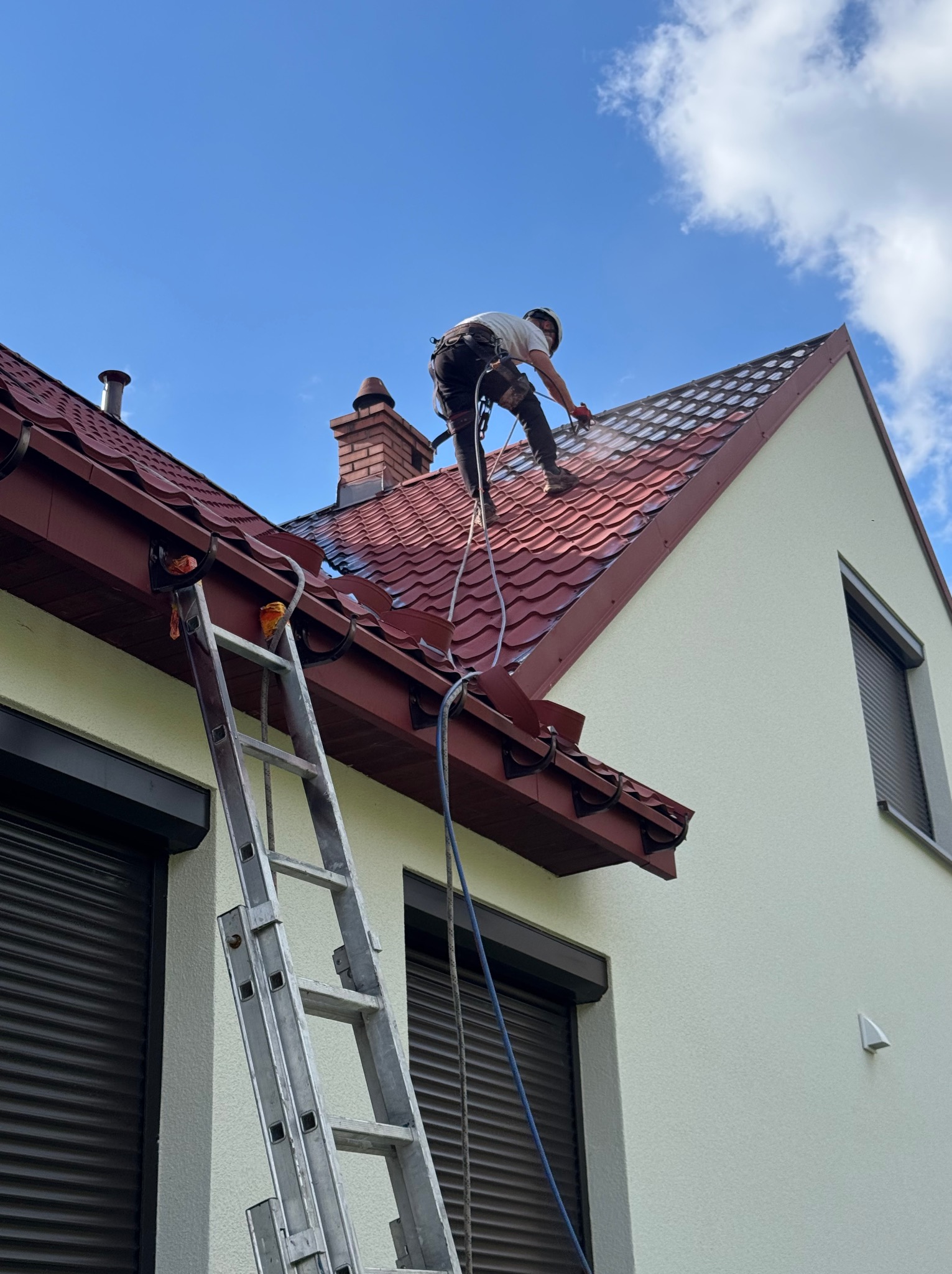 Pracownik w uprzęży, z drabiny, czyści czerwoną dachówkę domu jednorodzinnego za pomocą myjki ciśnieniowej. Widoczne zacieki i czyszczona powierzchnia.