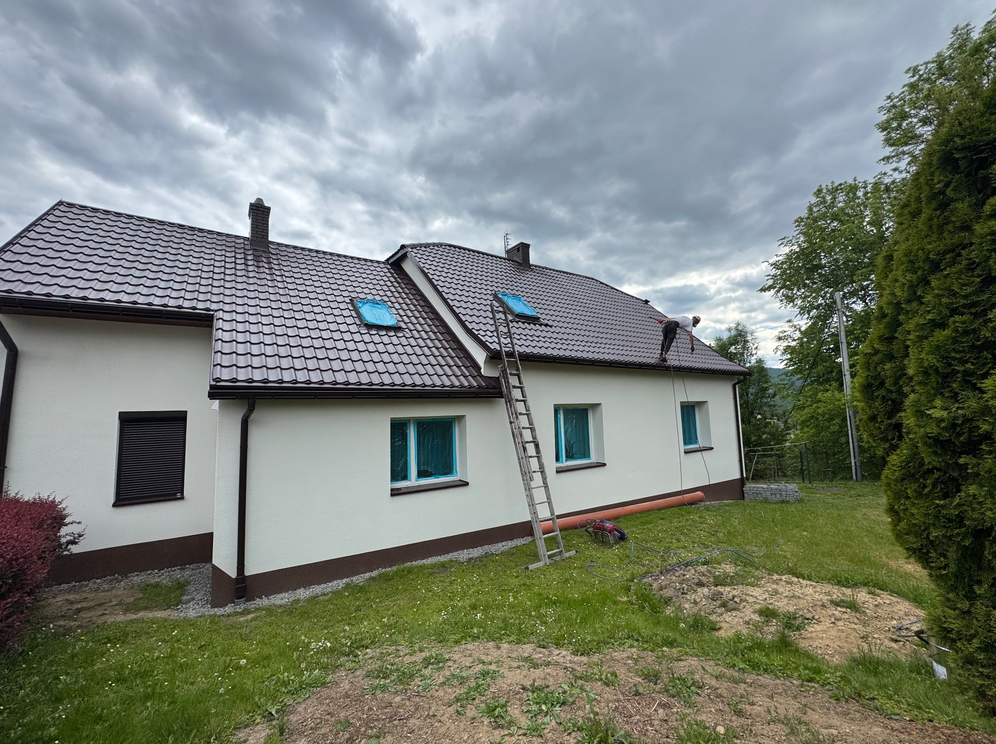 Dom jednorodzinny z brązowym dachem, zabezpieczonymi oknami i drabiną. Widoczny fragment elewacji po malowaniu. Pochmurne niebo w tle.