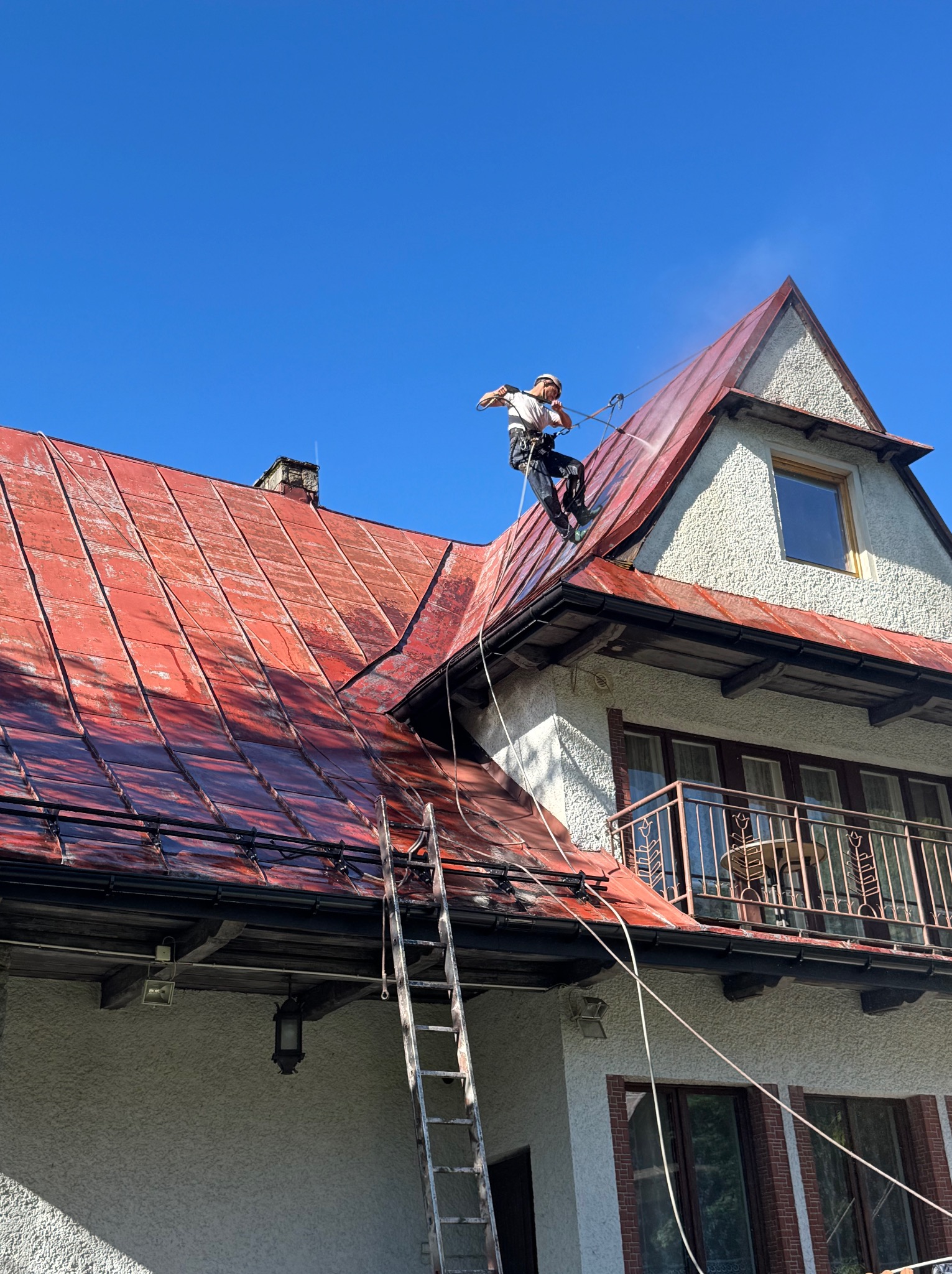 Pracownik w uprzęży, z drabiny, spryskuje wodą czerwoną dachówkę na dwuspadowym dachu domu z poddaszem, na tle błękitnego nieba. Widoczne zacieki.