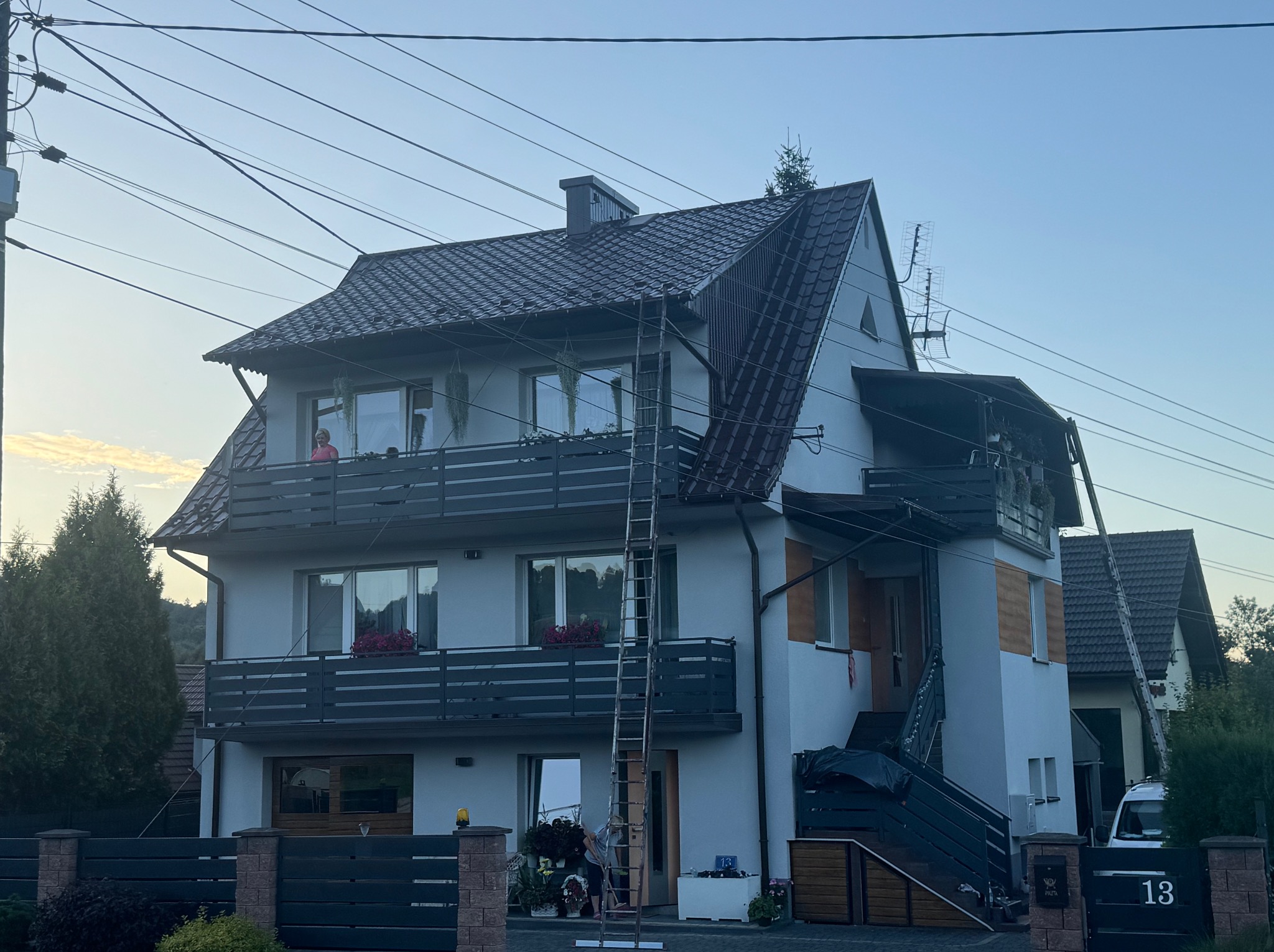 Dwupiętrowy dom z balkonami i drabiną opartą o elewację. Widoczna osoba na balkonie i przy drabinie. Ujęcie z dołu, widoczne przewody elektryczne.