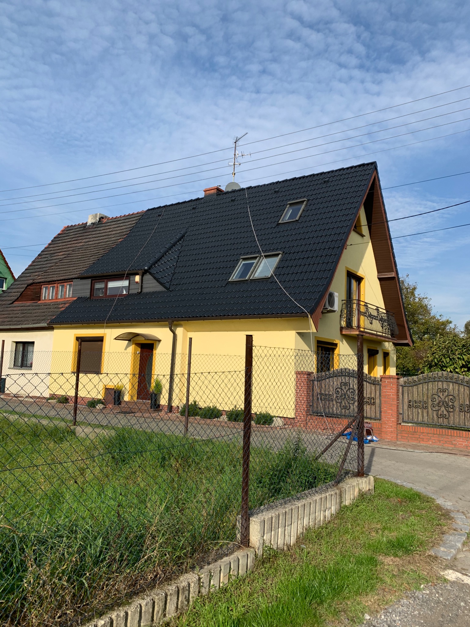 Żółty dom jednorodzinny z nowym, czarnym dachem z dachówki, widoczne elementy wykończeniowe i rynny, ogrodzony siatką, z fragmentem starego dachu po lewej stronie.