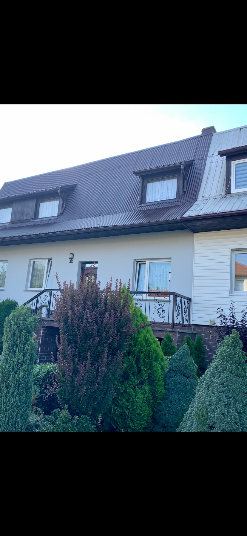 Dwukondygnacyjny dom z brązowym dachem z blachy trapezowej i jasną elewacją, ozdobiony zielenią w ogrodzie.