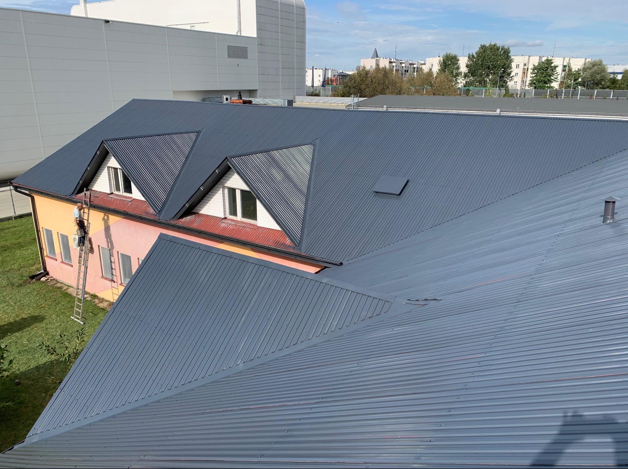 Blaszany dach pomalowany na szaro, widok z góry, pracownik na drabinie przy budynku z różową elewacją, w tle zabudowa miejska.