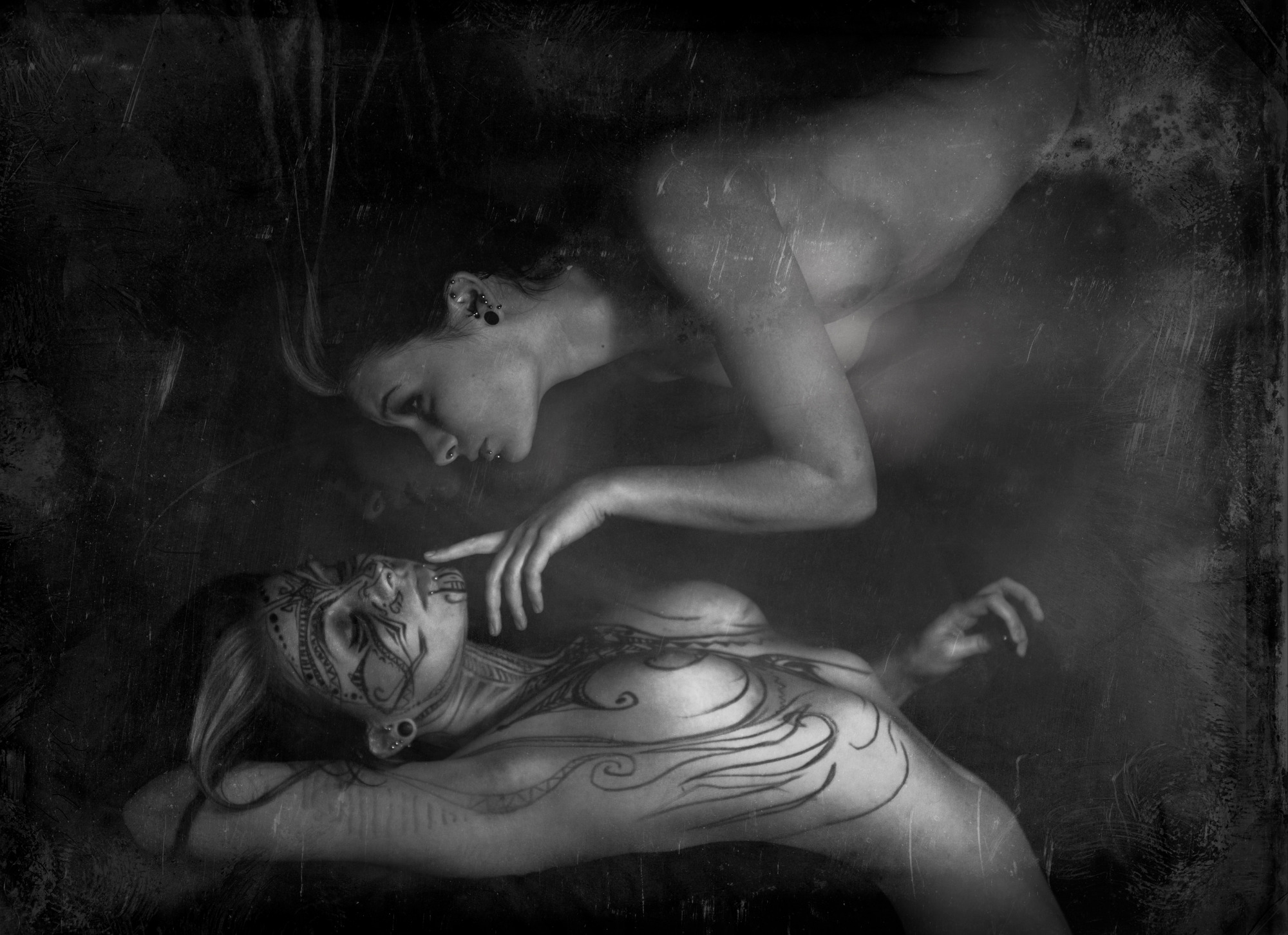 Czarno-biały fotomontaż przedstawiający dwie kobiety, jedna z tatuażami na twarzy i ciele leżąca na plecach, druga unosząca się nad nią, dotykająca jej twarzy.