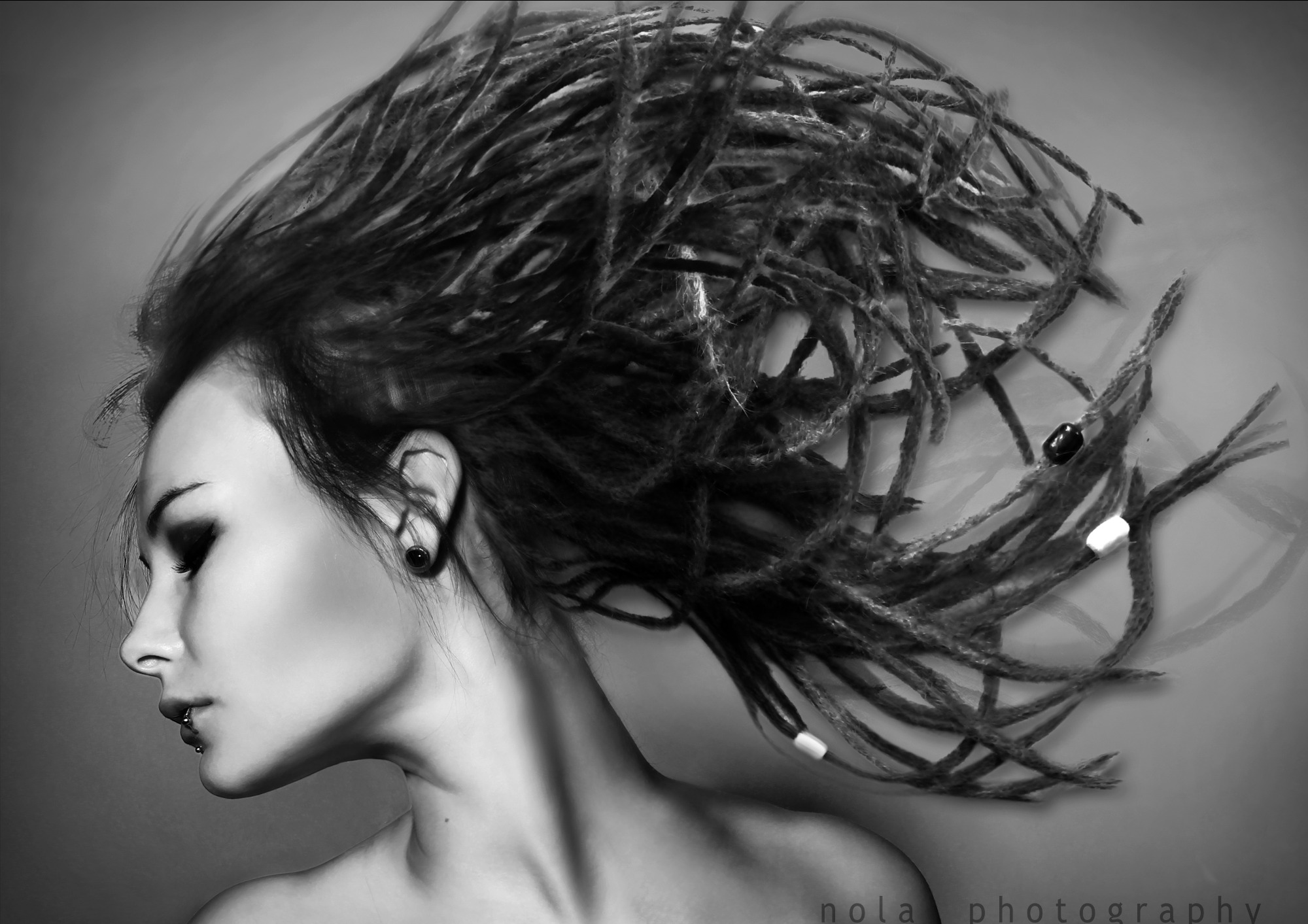 Czarno-biała fotografia studyjna profilu kobiety z dredami w ruchu, z widocznym kolczykiem w uchu i piercingiem w wardze, na gładkim tle.