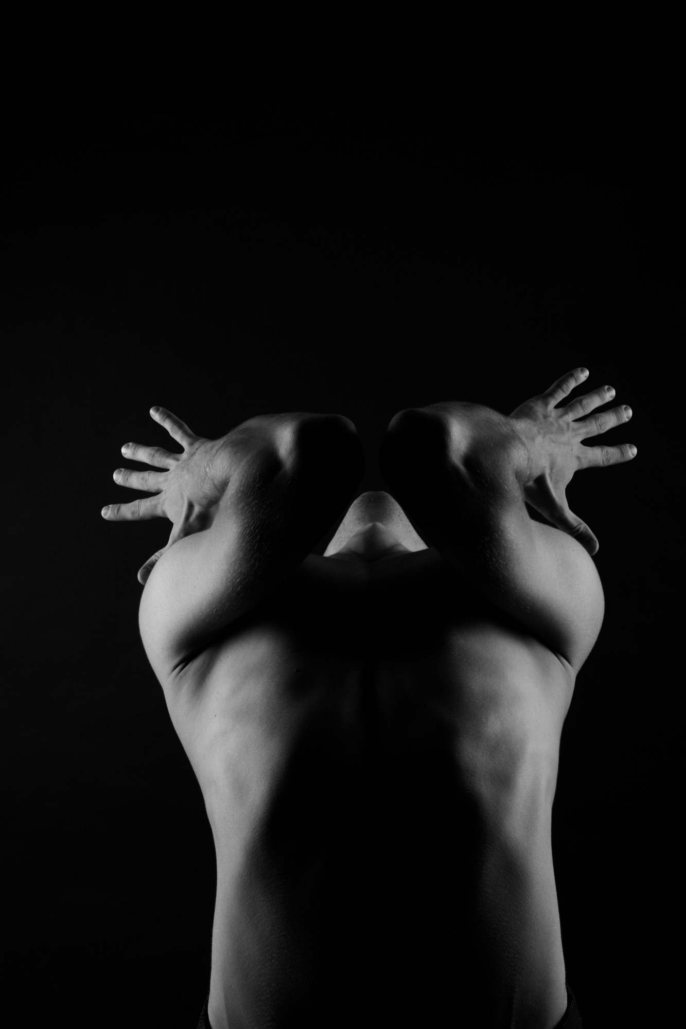 Czarno-biała fotografia studyjna, przedstawiająca tors i ramiona umięśnionego mężczyzny, z dłońmi uniesionymi nad głową na ciemnym tle.