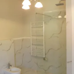 Nowoczesna łazienka z marmurowymi płytkami, szklanym panelem prysznicowym, grzejnikiem drabinkowym, bidetem i designerską lampą sufitową.