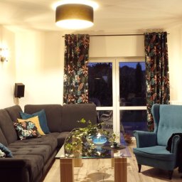 Jarońska Wnętrza projektowanie wnętrz - Salon z narożną sofą, turkusowym fotelem i ścianą z cegły, oświetlony lampą sufitową i wiszącą, z roślinami doniczkowymi i dekoracjami na szklanym stoliku kawowym.