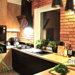 Jarońska Wnętrza projektowanie wnętrz - Nowoczesna kuchnia z drewnianymi szafkami, czarnymi frontami i ceglaną ścianą, oświetlona lampami wiszącymi, z roślinami doniczkowymi i koszem pełnym warzyw na blacie.
