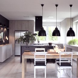 Jarońska Wnętrza projektowanie wnętrz - Jasna kuchnia z drewnianym stołem i białymi krzesłami, nad którymi wiszą trzy czarne lampy w stylu industrialnym, z przyległą wyspą kuchenną i zabudową w odcieniach szarości oraz czarna ściana...