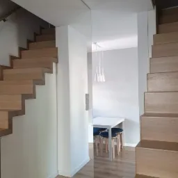 Nowoczesne drewniane schody dywanowe z widokiem na minimalistyczną jadalnię z białym stołem i niebieskimi krzesłami, oświetloną trzema wiszącymi lampami.