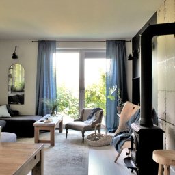 Jarońska Wnętrza projektowanie wnętrz - Jasne wnętrze salonu z ciemnoszarą sofą narożną, drewnianym stołem kawowym, fotelem w stylu retro, piecem wolnostojącym i regałem z książkami, doświetlone naturalnym światłem przez okno balkonowe...