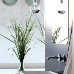 Jarońska Wnętrza projektowanie wnętrz - Nowoczesna łazienka z białą, prostokątną umywalką, chromowaną baterią, wiszącymi żarówkami dekoracyjnymi i trawą w szklanym wazonie na tle okrągłych okien.