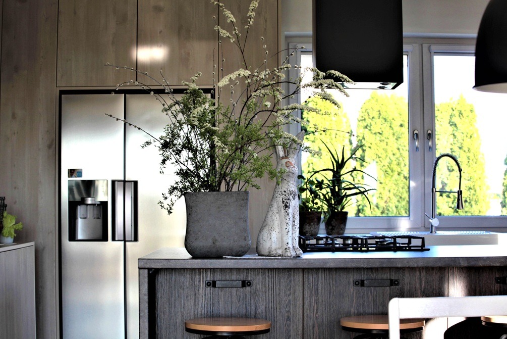 Nowoczesna kuchnia z wyspą, na której stoi betonowa donica z kwiatami, ceramiczny królik i roślina doniczkowa, w tle okno z widokiem na zieleń, po lewej lodówka side-by-side.