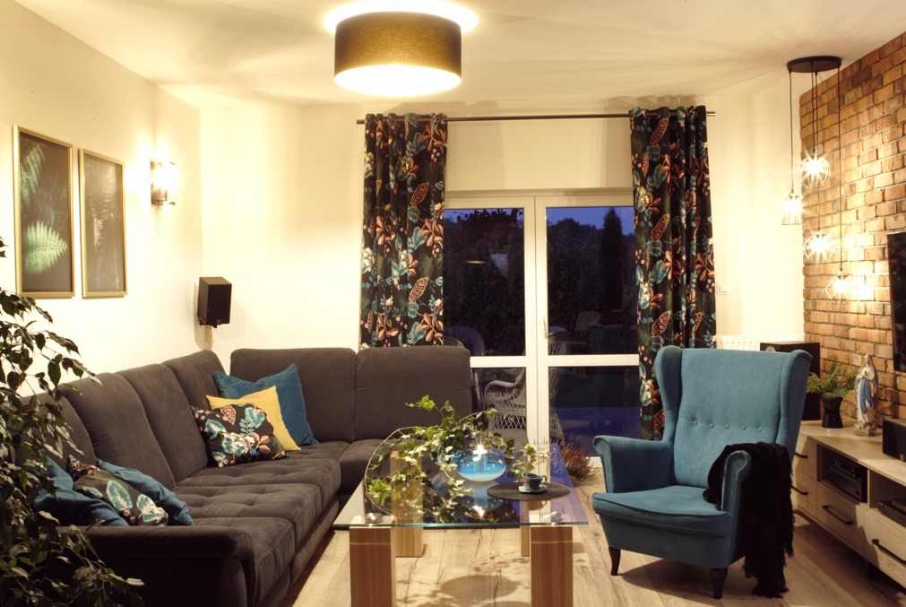 Salon z narożną sofą, turkusowym fotelem i ścianą z cegły, oświetlony lampą sufitową i wiszącą, z roślinami doniczkowymi i dekoracjami na szklanym stoliku kawowym.