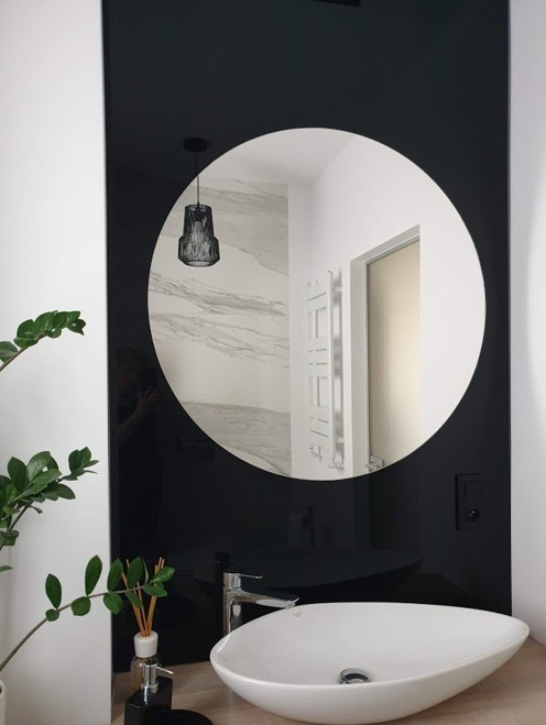 Nowoczesna łazienka z okrągłym lustrem w czarnej ramie, białą umywalką w kształcie łzy i minimalistyczną armaturą, uzupełniona o czarny dyfuzor zapachowy i zieloną roślinę doniczkową.