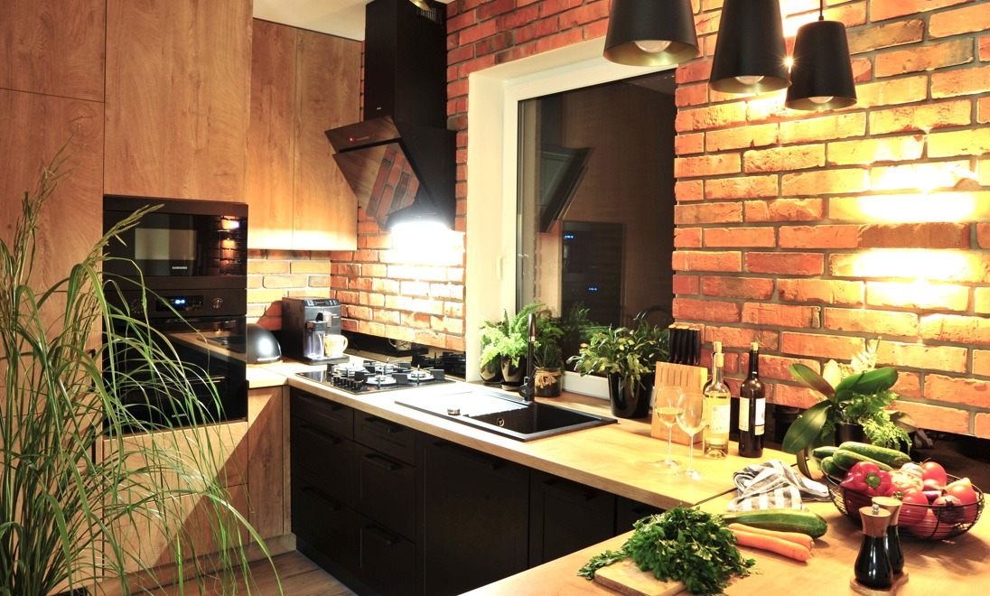 Nowoczesna kuchnia z drewnianymi szafkami, czarnymi frontami i ceglaną ścianą, oświetlona lampami wiszącymi, z roślinami doniczkowymi i koszem pełnym warzyw na blacie.