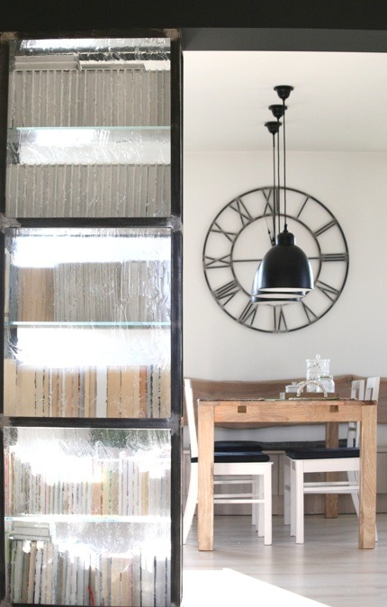 Przeszklona ścianka działowa z czarnymi ramami, oddzielająca przestrzeń jadalną z drewnianym stołem, białymi krzesłami i wiszącą lampą na tle ściany z dużym zegarem.