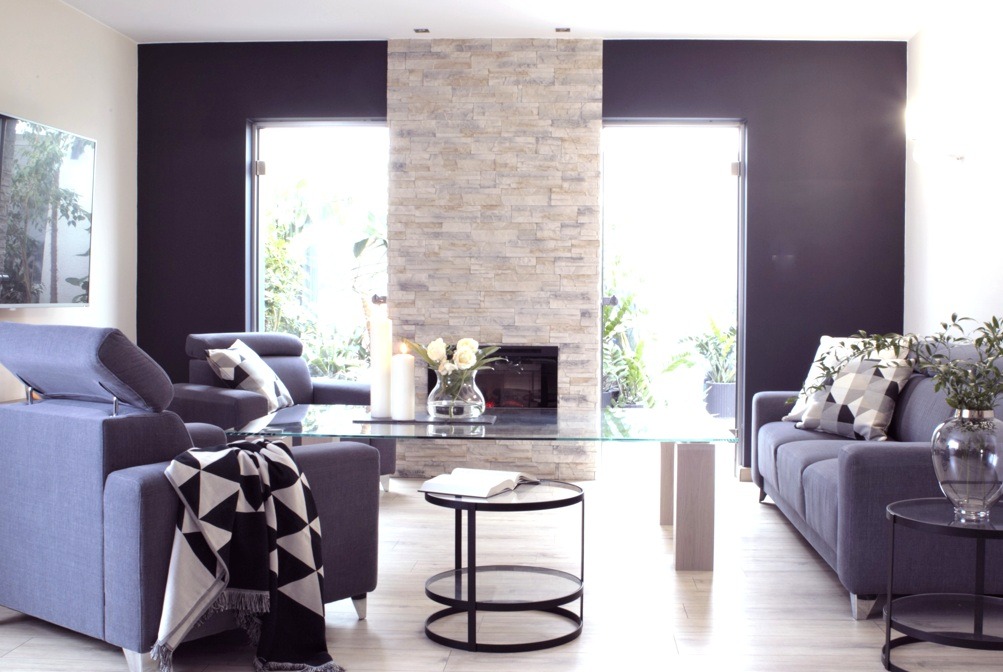 Nowoczesny salon z szarymi sofami, szklanym stołem, kominkiem z kamienia dekoracyjnego, czarną ścianą i dużymi oknami z widokiem na zieleń.