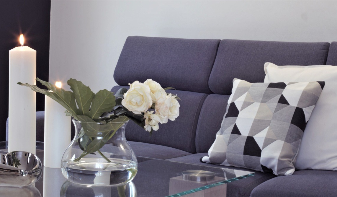 Stylowe wnętrze: bukiet białych róż w szklanym wazonie, zapalone świece i poduszki geometryczne na szarej sofie.
