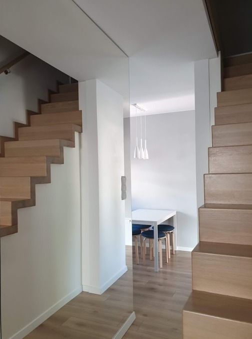 Nowoczesne drewniane schody dywanowe z widokiem na minimalistyczną jadalnię z białym stołem i niebieskimi krzesłami, oświetloną trzema wiszącymi lampami.