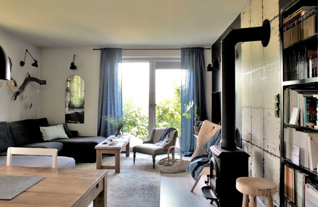 Jasne wnętrze salonu z ciemnoszarą sofą narożną, drewnianym stołem kawowym, fotelem w stylu retro, piecem wolnostojącym i regałem z książkami, doświetlone naturalnym światłem przez okno balkonowe...